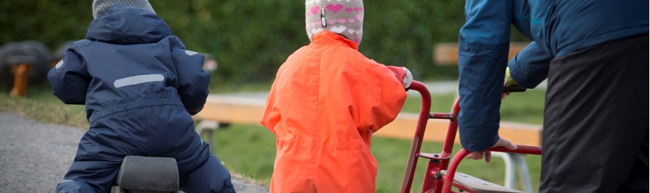 Børn i flyverdragter kører på løbecykel udenfor på legeplads sammen med pædagogisk assistent