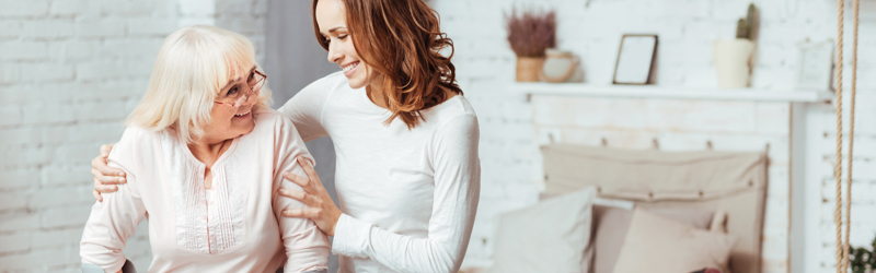 Billedet viser en yngre kvinde, der hjælper en ældre kvinde med krykker.